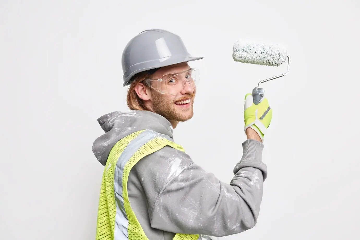 A man painting a new home, holding a roller, wearing a protective helmet, glasses, and a uniform, doing repair work, posing against a white background.