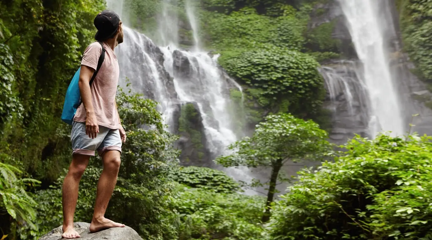 A young barefoot tourist wearing a baseball cap stands on a large stone, looking back at the waterfall behind him in a beautiful, exotic natural setting. The bearded traveler enjoys the wildlife while hiking through the rainforest.